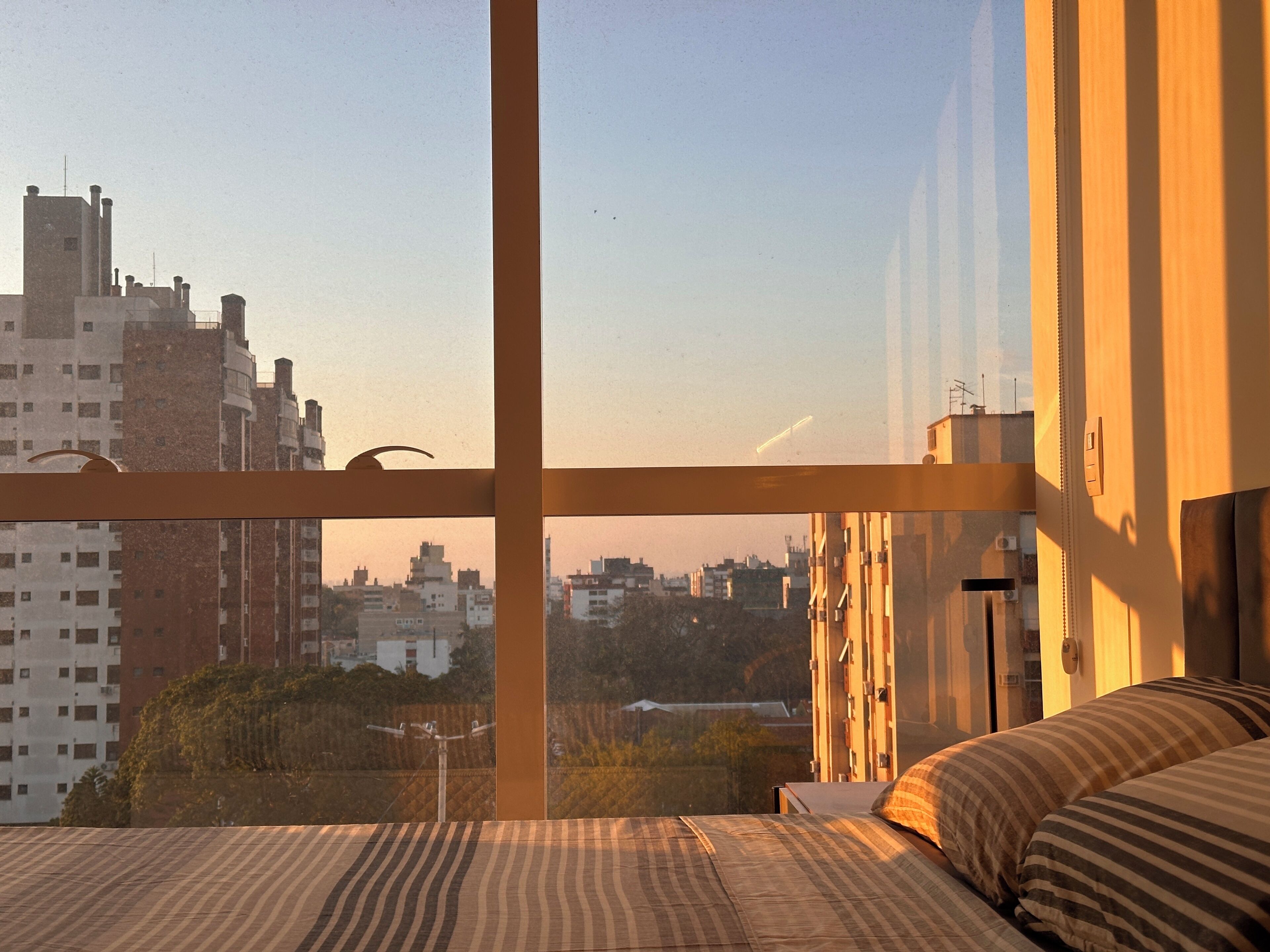 A hotel room with a large window, a bed with striped bedding, and a view of the cityscape.