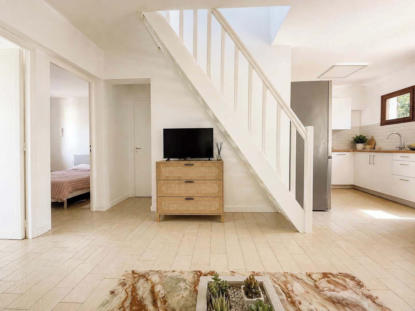 A modern interior with a staircase leading to a loft bedroom, a kitchen with white cabinets, and a flat-screen TV on a wooden dresser.