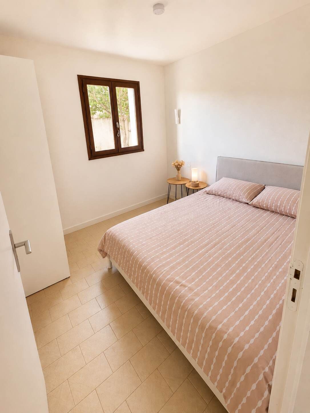 Ein Schlafzimmer mit einem Bett, einem Fenster, einem Nachttisch mit Lampe und einer Tür.