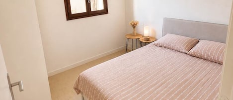 Un dormitorio con cama, ventana, mesita de noche con lámpara y una puerta.