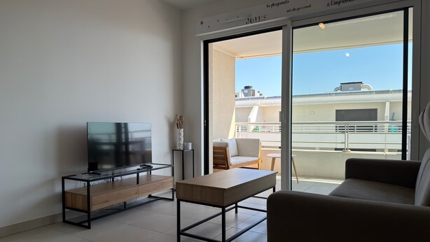 A modern living room with a flat-screen TV, a wooden coffee table, a sofa, and a balcony with a view of the city. - Valras- Loc Archipel Panarea  vue mer (Valras-Plage)