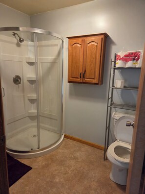 A bathroom with a glass shower enclosure, a toilet, and wooden cabinets above the toilet. - Cozy loft apartment in historic downtown Cole Camp. (Cole Camp)