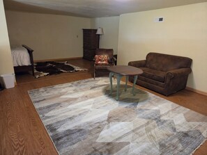 Ein Wohnzimmer mit einem braunen Sofa, einem kleinen Tisch, einem gemusterten Teppich, Holzboden, einem Fellteppich und einer dunklen Holzkommode.