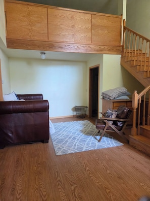 Ein Loft-Zimmer mit Holzbalkendecke, einem braunen Leder-Sofa, einem gemusterten Teppich, einem kleinen Tisch mit Stuhl und einer Treppe.