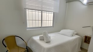 A single bed with a white mattress and pillows, a chair with a yellow seat, and a window with blinds. - Rodadero Relax | Walk to Beach & Park (Santa Marta)