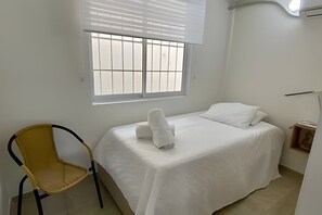 Ein Einzelbett mit weißer Matratze und Kissen, ein Stuhl mit gelber Sitzfläche und ein Fenster mit Jalousien.
