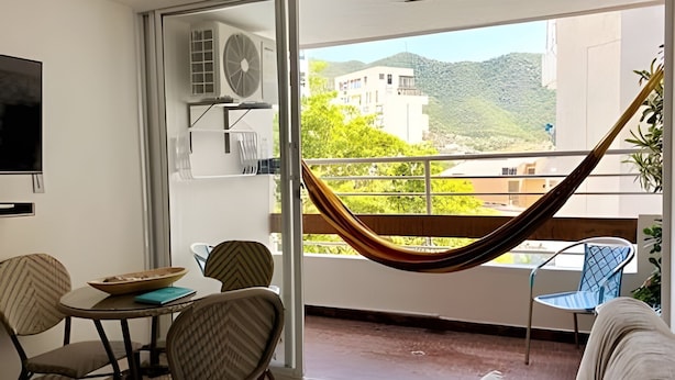 A balcony with a hammock, a dining table with chairs, and a view of buildings and greenery. - Rodadero Relax | Walk to Beach & Park (Santa Marta)