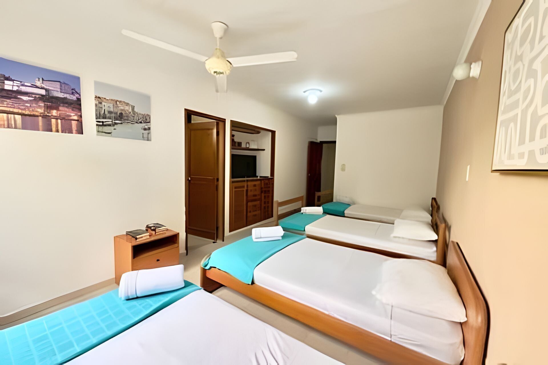 A hotel room with three beds, a ceiling fan, a small table with books, and a wardrobe.