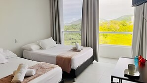 A hotel room with two beds, a large window with a view of greenery, and a small table with a cup and decorative items. - Bello Horizonte Haven | Resort Pools & Beach (Santa Marta)