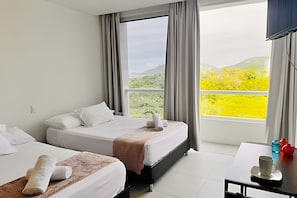 Quarto de hotel com duas camas, uma janela ampla com vista para o verde e uma pequena mesa com uma xícara e objetos decorativos.