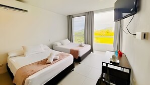A hotel room with two beds, a flat-screen TV mounted on the wall, and a window with a view of greenery.
