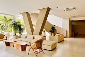 Um lobby moderno com um elemento arquitetônico em formato de V, área de estar com sofás e poltronas, e plantas em vasos.