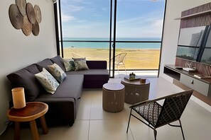 A modern living room with a large window overlooking a beach, a sofa, a coffee table, and a TV. - Ocean view, swimming pools, gym, and beach access (Santa Marta)