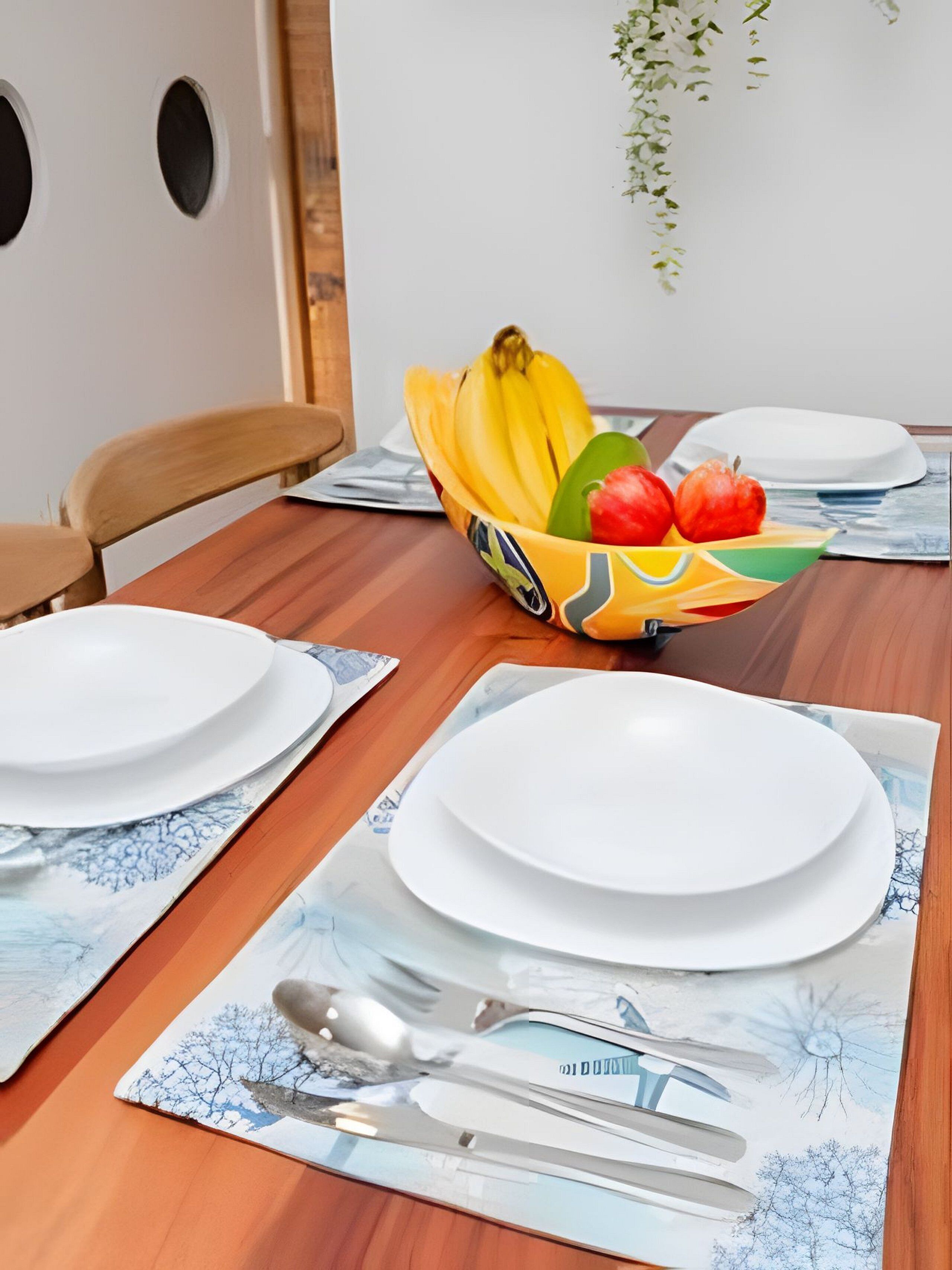 A dining table set with white plates, a bowl of fruit, and placemats with a tree design.