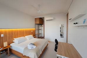 A modern hotel room with a wooden headboard, a bed with white linens, a desk with a chair, and a wardrobe.