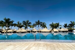 Une piscine bordée de palmiers et entourée de constructions aux toits de chaume.