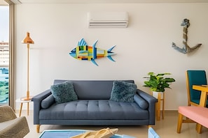 Un salon avec un canapé gris, deux coussins décoratifs, une décoration murale représentant des poissons aux couleurs vives, une plante en pot et une chaise en bois.