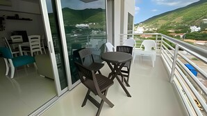 En balkon med et bord og stole, en glasdør der fører til et rum, og udsigt over et grønt landskab med bakker.