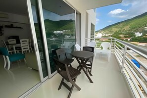 A balcony with a table and chairs, a glass door leading to a room, and a view of a green landscape with hills. - Rodadero Escape | Pool, Balcony, BBQ & Parking (Santa Marta)
