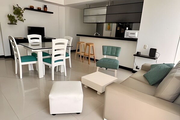 A modern living room with a white sofa, a white coffee table, and a dining area with white chairs and a glass table. - Rodadero Escape | Pool, Balcony, BBQ & Parking (Santa Marta)