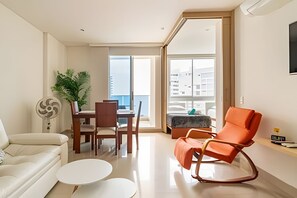 Uma sala de estar moderna com um sofá, uma mesa de jantar, cadeiras, uma mesa de centro e uma poltrona de balanço. Há uma janela grande que oferece vista para edifícios.