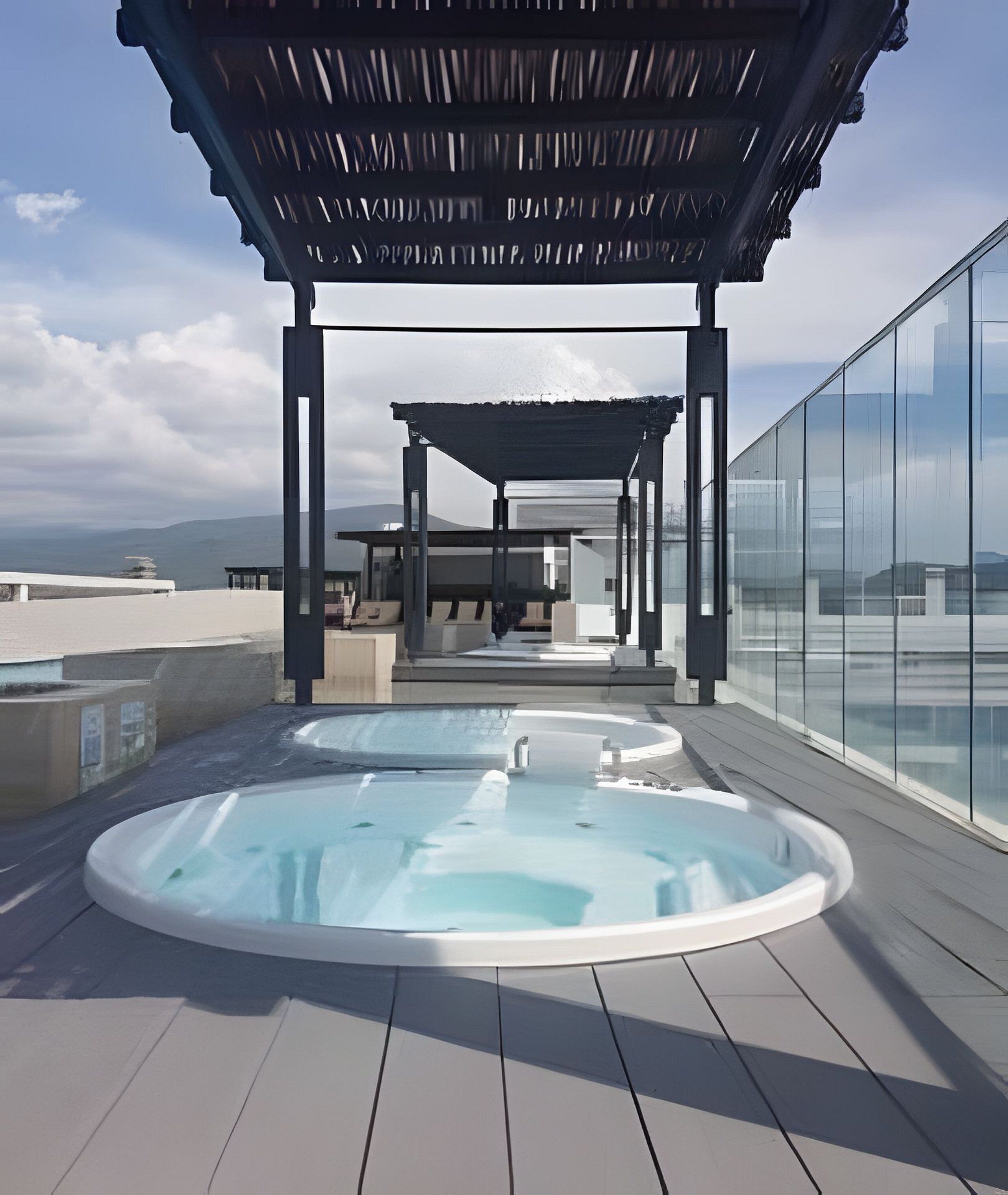 A rooftop terrace with two hot tubs, a covered seating area, and a glass wall offering a city view.