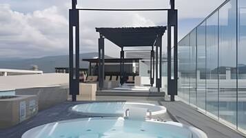 A rooftop terrace with two hot tubs, a covered seating area, and a glass wall offering a city view.