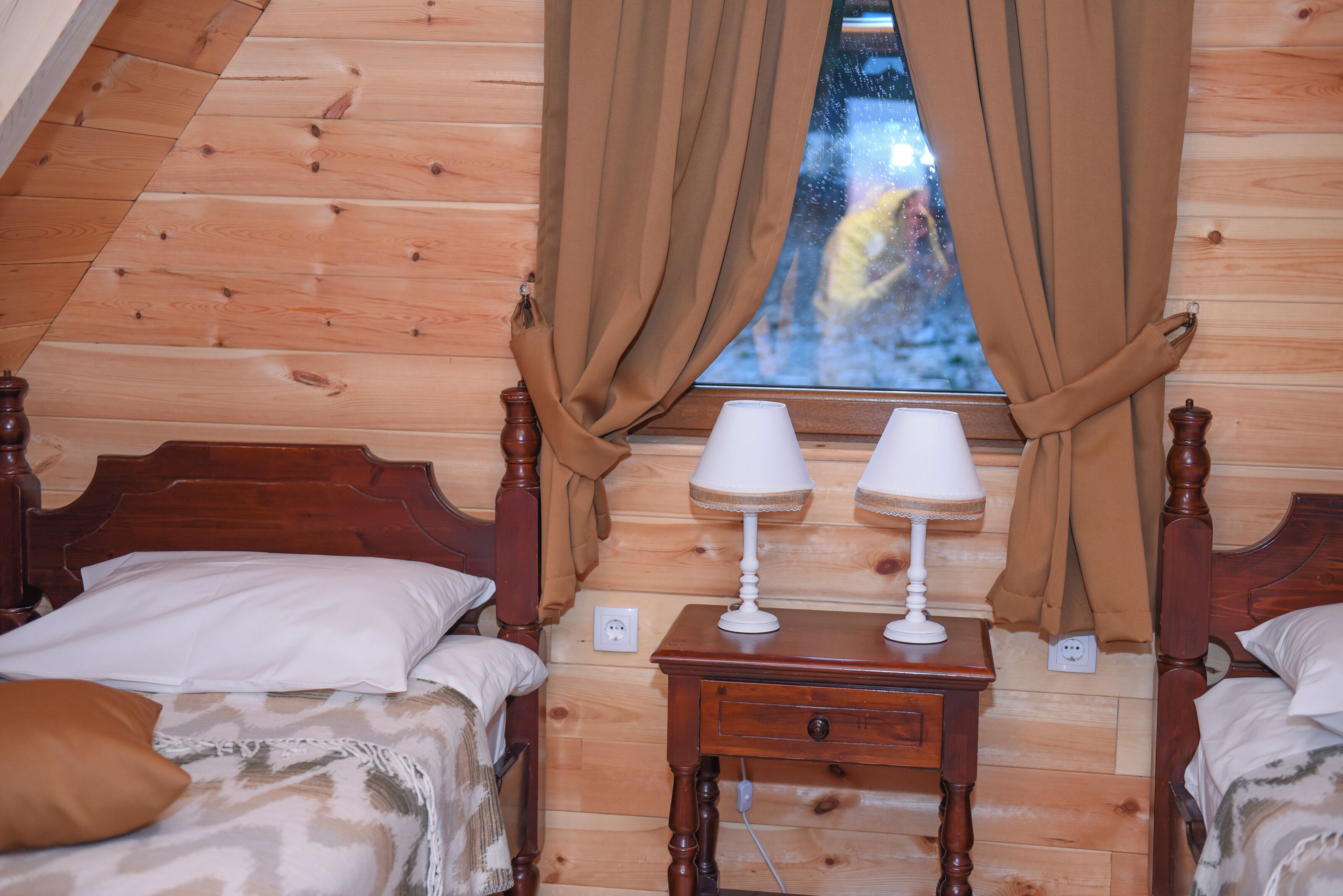 Een gezellige kamer met houten wanden en een raam met gordijnen. Er staan twee bedden met kussens en een klein houten nachtkastje met twee lampen.