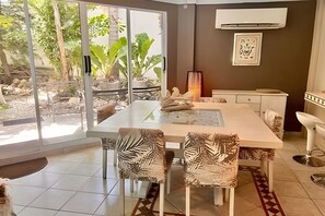 A modern dining area with a glass door leading to an outdoor garden, a white table with chairs, and a small kitchen area. - Lagomar House: Private Pool, BBQ & Beach (Santa Marta)