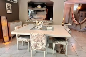 A dining area with a large table and chairs, a kitchen in the background, and a hammock in the corner.