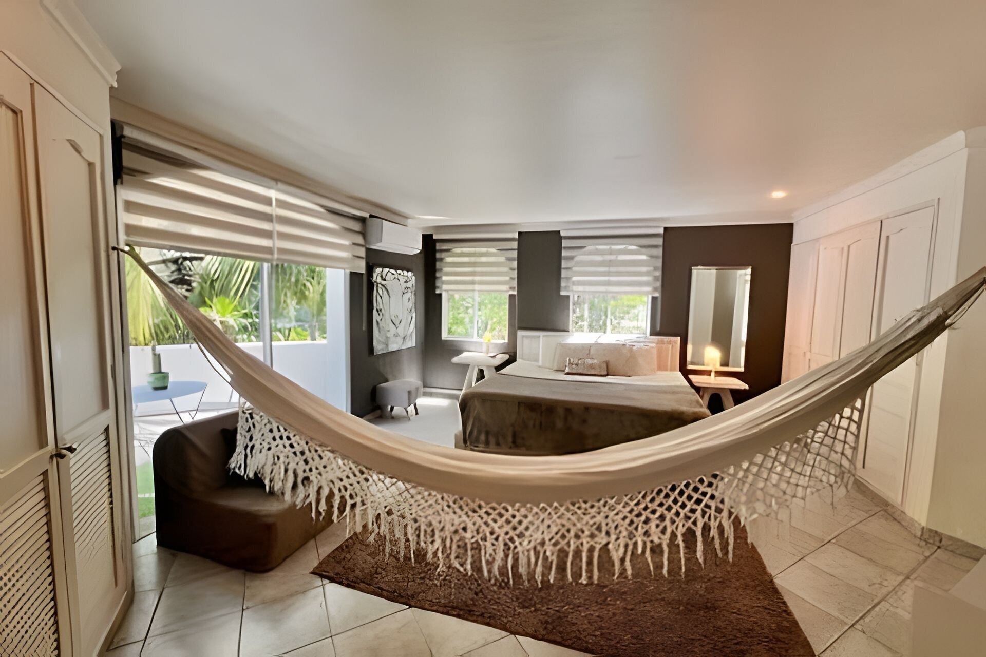 A bedroom with a hammock, a bed, a small table, a chair, and a large window.