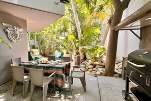 An outdoor dining area with a table and chairs, surrounded by greenery and a barbecue grill.