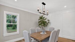 A dining room with a wooden table, six grey chairs, a chandelier, and a potted plant. - 7BR, 6.5Bath NEW Construction Family Home in Barnstable Village (Barnstable)