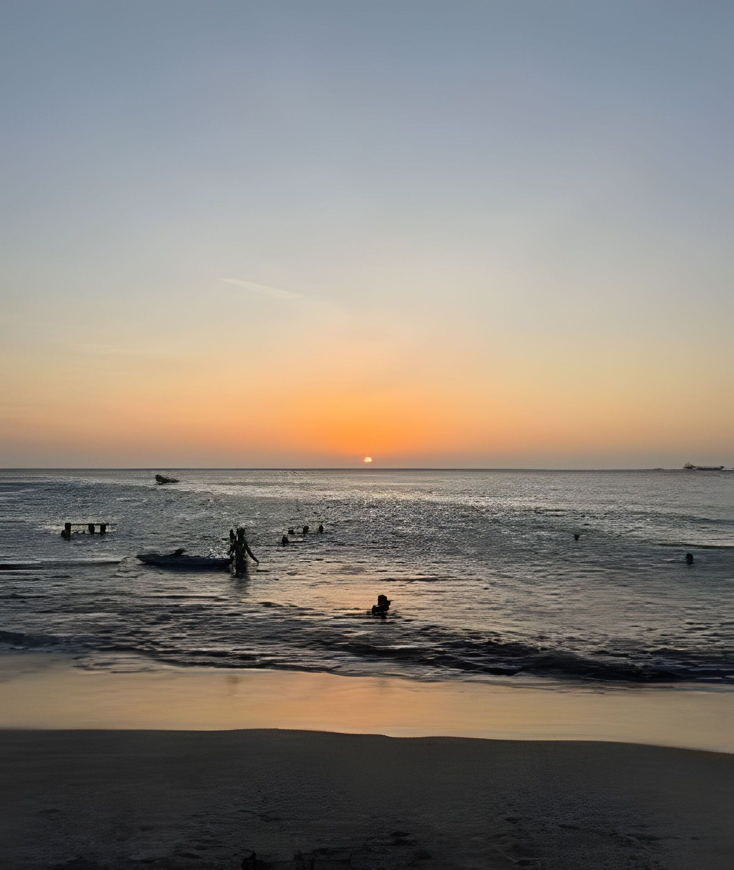 A beach scene with the sun setting over the ocean, people swimming, and a ship in the distance.