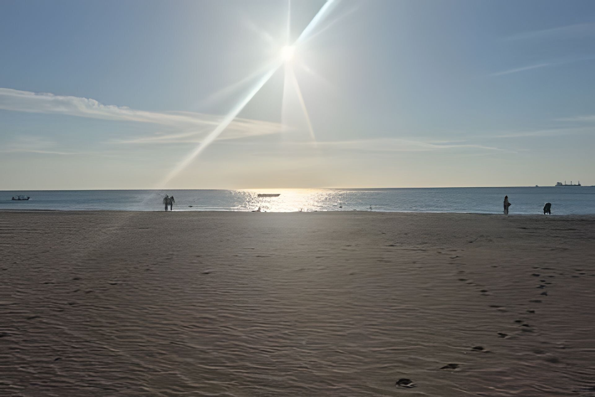 Une plage où l’on voit des empreintes de pas menant à l’eau, un bateau au loin et un ciel dégagé.