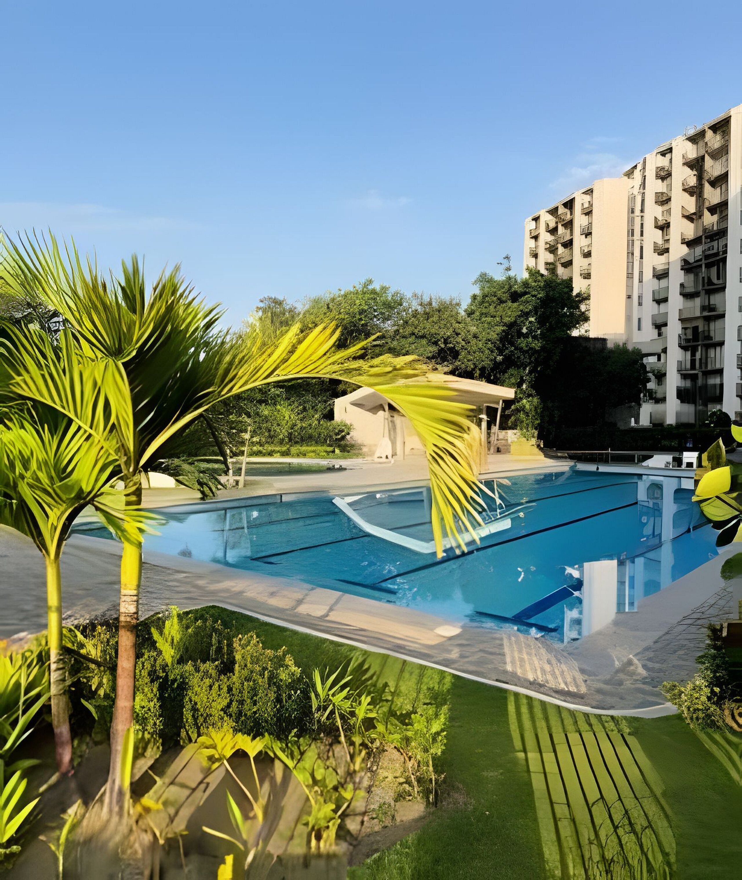 Une piscine entourée d’un jardin, avec un immeuble à plusieurs étages en arrière-plan.