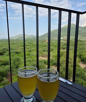 Two glasses of yellow beverage on a table with a view of green hills and mountains.