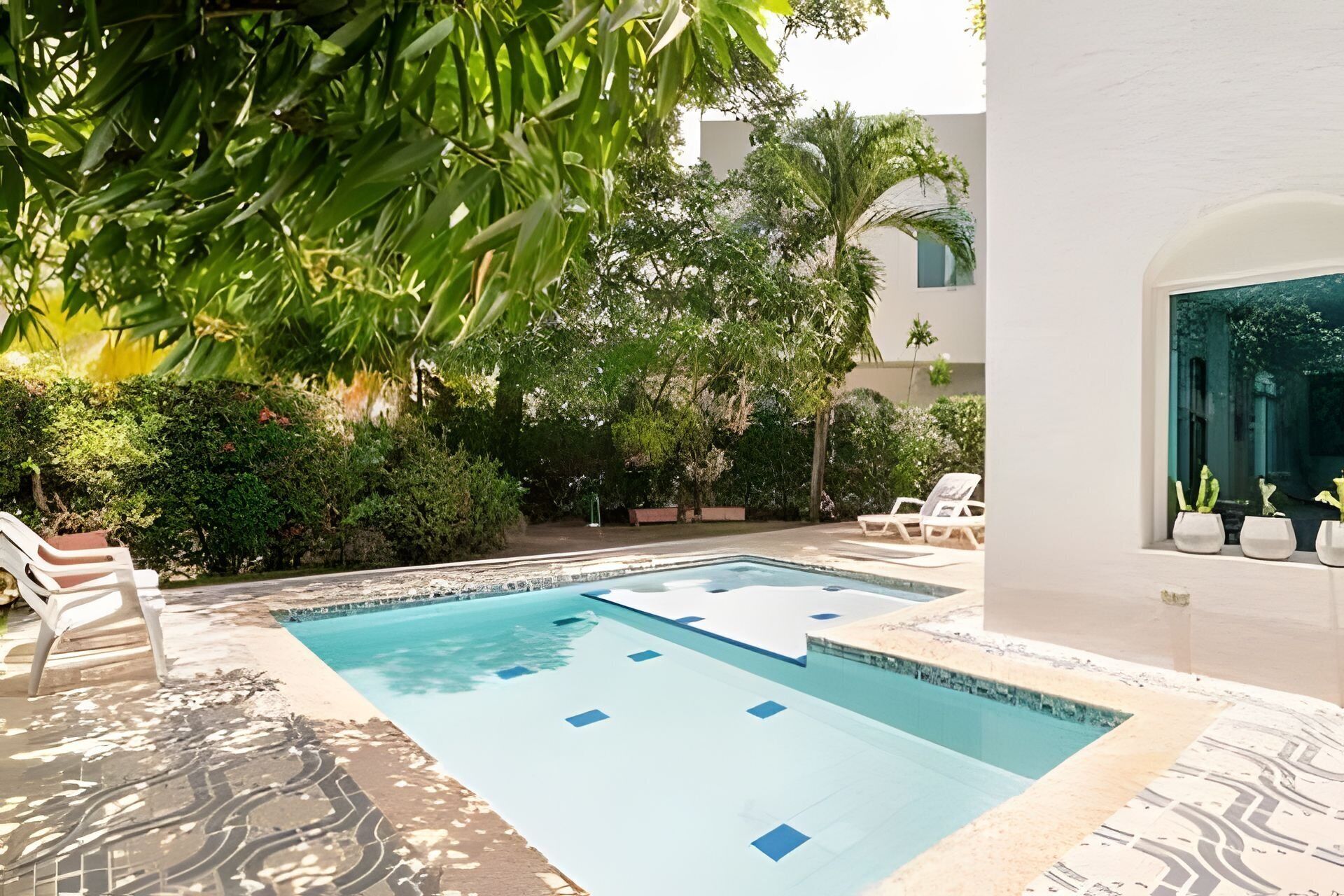 Une piscine dotée d’une terrasse carrelée, entourée de verdure et bordée d’un bâtiment blanc.