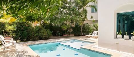 A swimming pool with a tiled deck, surrounded by greenery and a white building.