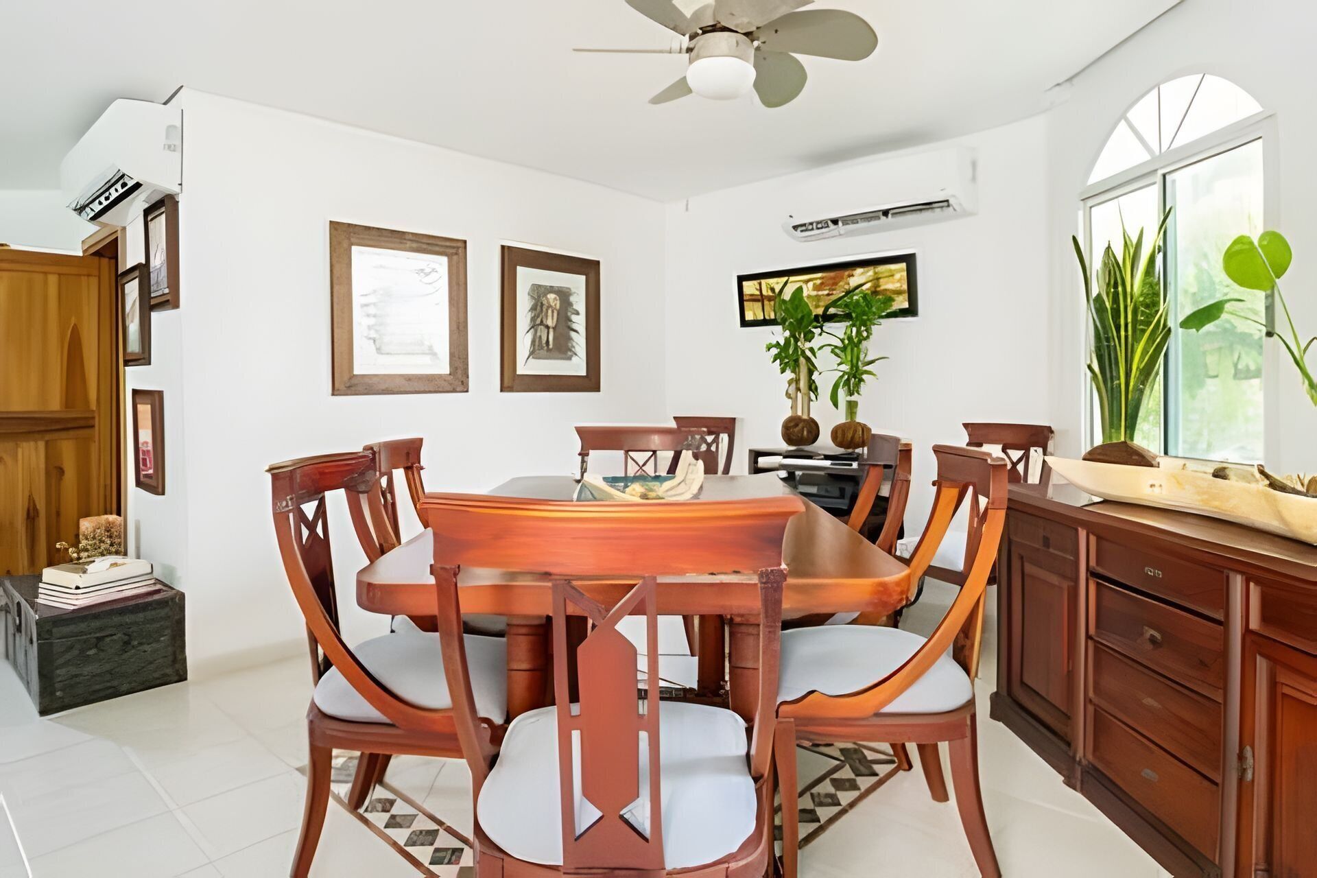 A dining area with a wooden table and chairs, framed artwork on the walls, and a large window with plants.