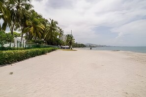 Ein weiter, sandiger Strand mit blauem Himmel, Palmen und einer Stadtlandschaft in der Ferne.