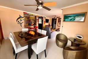 Un coin repas avec une table à plateau en verre, des chaises blanches, un ventilateur de plafond et un tableau accroché au mur.