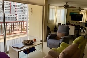 Ein Wohnzimmer mit einer Couch, Sesseln und einem Couchtisch. Es gibt einen Balkon mit Blick auf ein Gebäude.