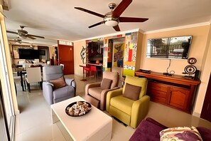 Ein Wohnzimmer mit Deckenventilator, ein Essbereich mit Fernseher und eine Küche im Hintergrund.