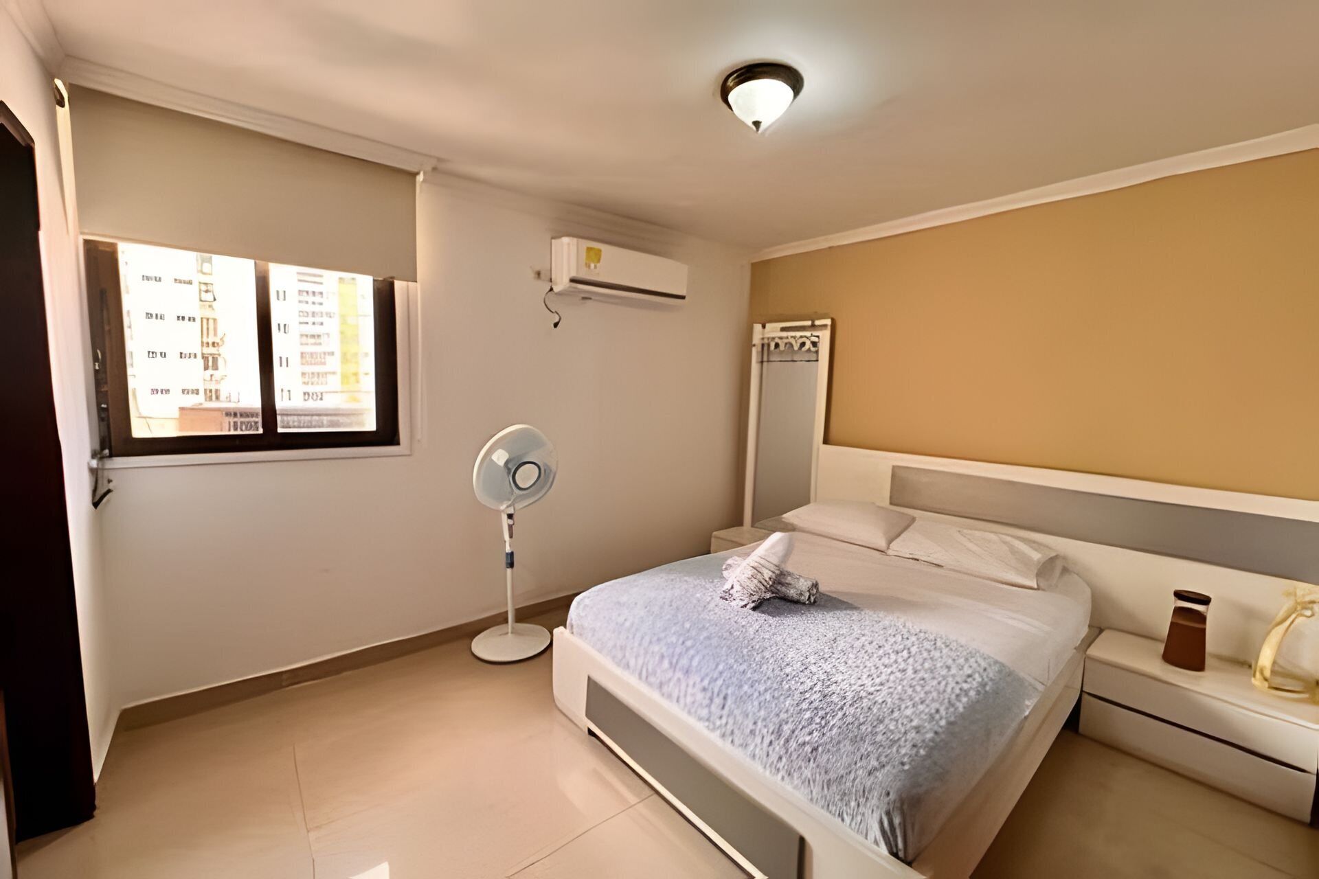 A bedroom with a bed, bedside table, nightstand, window, air conditioning unit, and a standing fan.