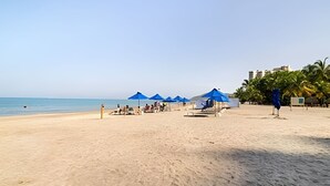 Ein Strand mit blauen Sonnenschirmen, einigen Personen und einer Pfauenstatue.