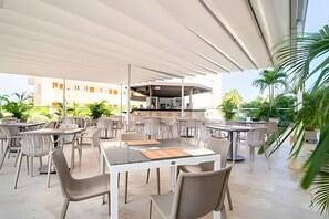 Ein Außenbereich mit Terrasse, Tischen, Stühlen, einer Bar und Blick auf ein Gebäude im Hintergrund.