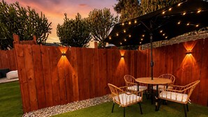 A patio area with a wooden fence, a round table with chairs, and string lights. - Private Patio & Fire Pit Near San Diego (Spring Valley)