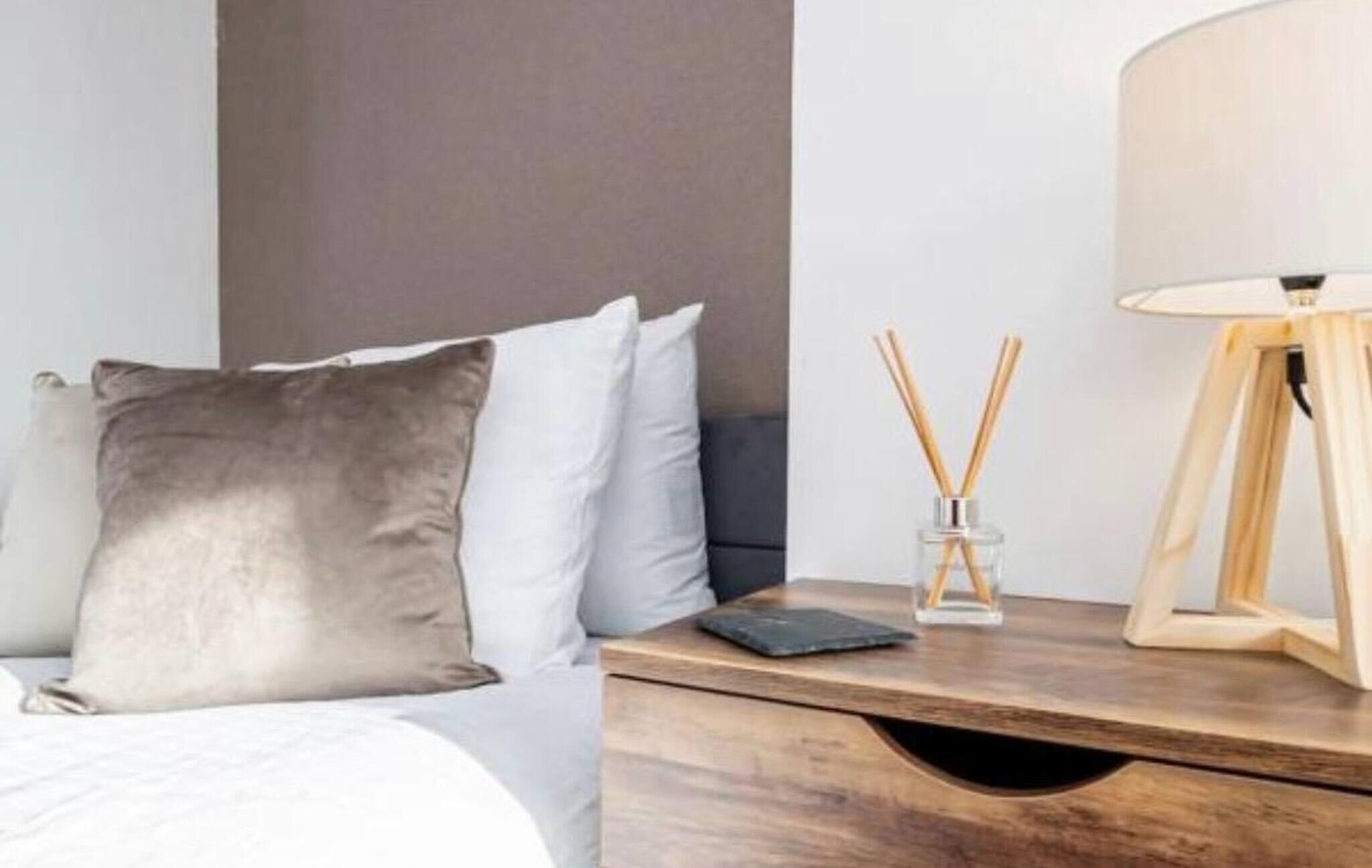 Een net opgemaakt bed met een grijs kussen, een nachtkastje met een lamp en een klein glazen vaasje met rietstokjes.
