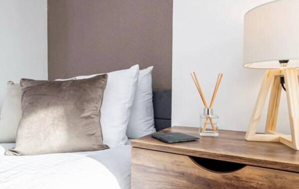A neatly made bed with a grey pillow, a bedside table with a lamp, and a small glass container with reed diffusers. - Stylish 3BR Home Near Nec, Bhx, HS2 & JLR (Birmingham)